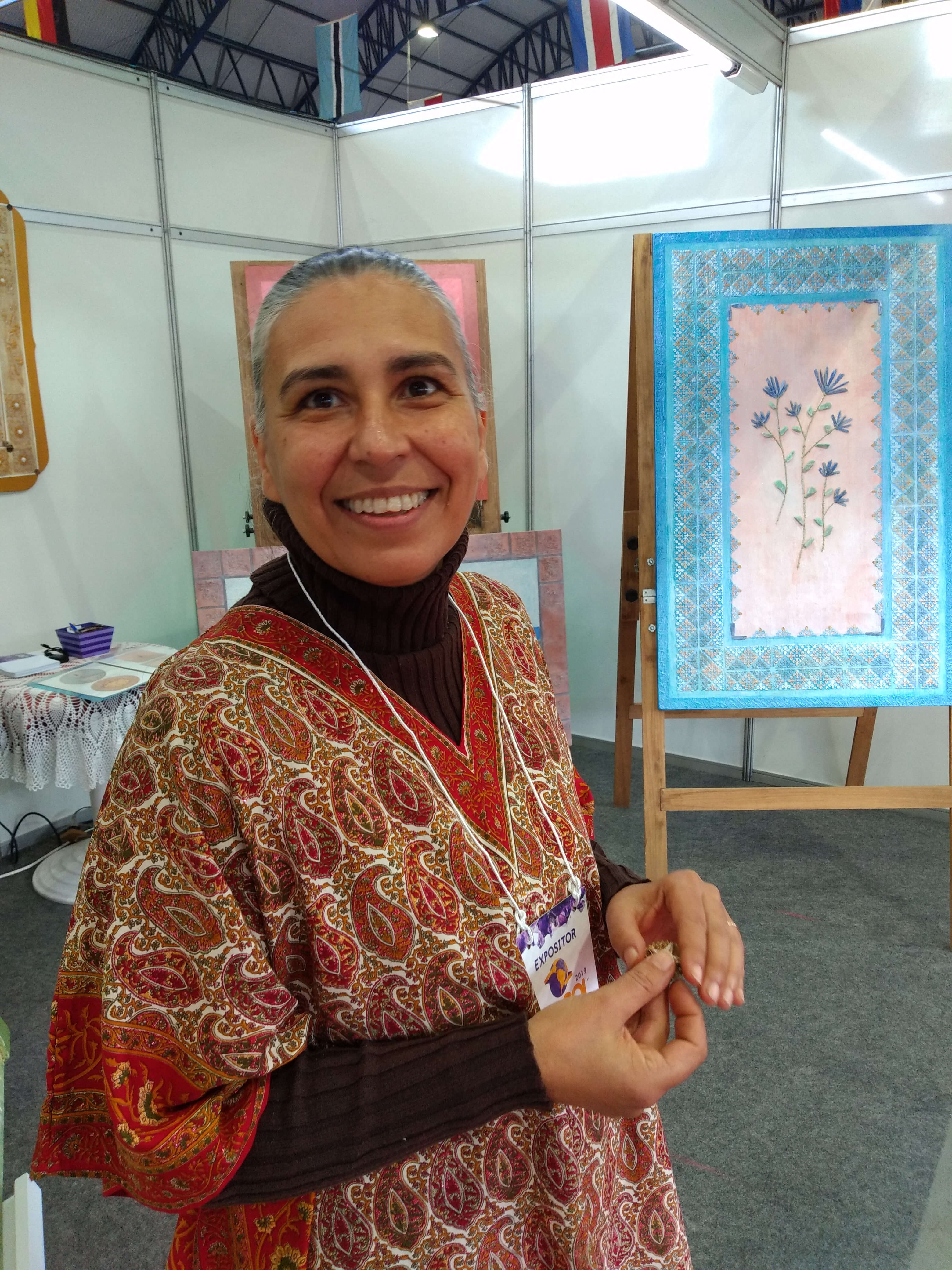 Isabel Arnold sorrindo para a foto em sua estande na Exposolo 2019 com suas obras ao fundo
